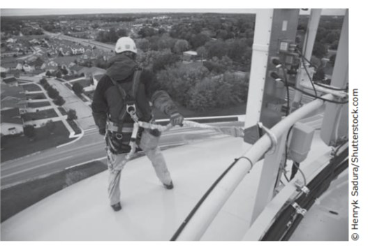 Photograph of a worker in safety gear standing on top of a tall water tower high above a neighborhood. Credit: Henryk Sadura/Shutterstock.com